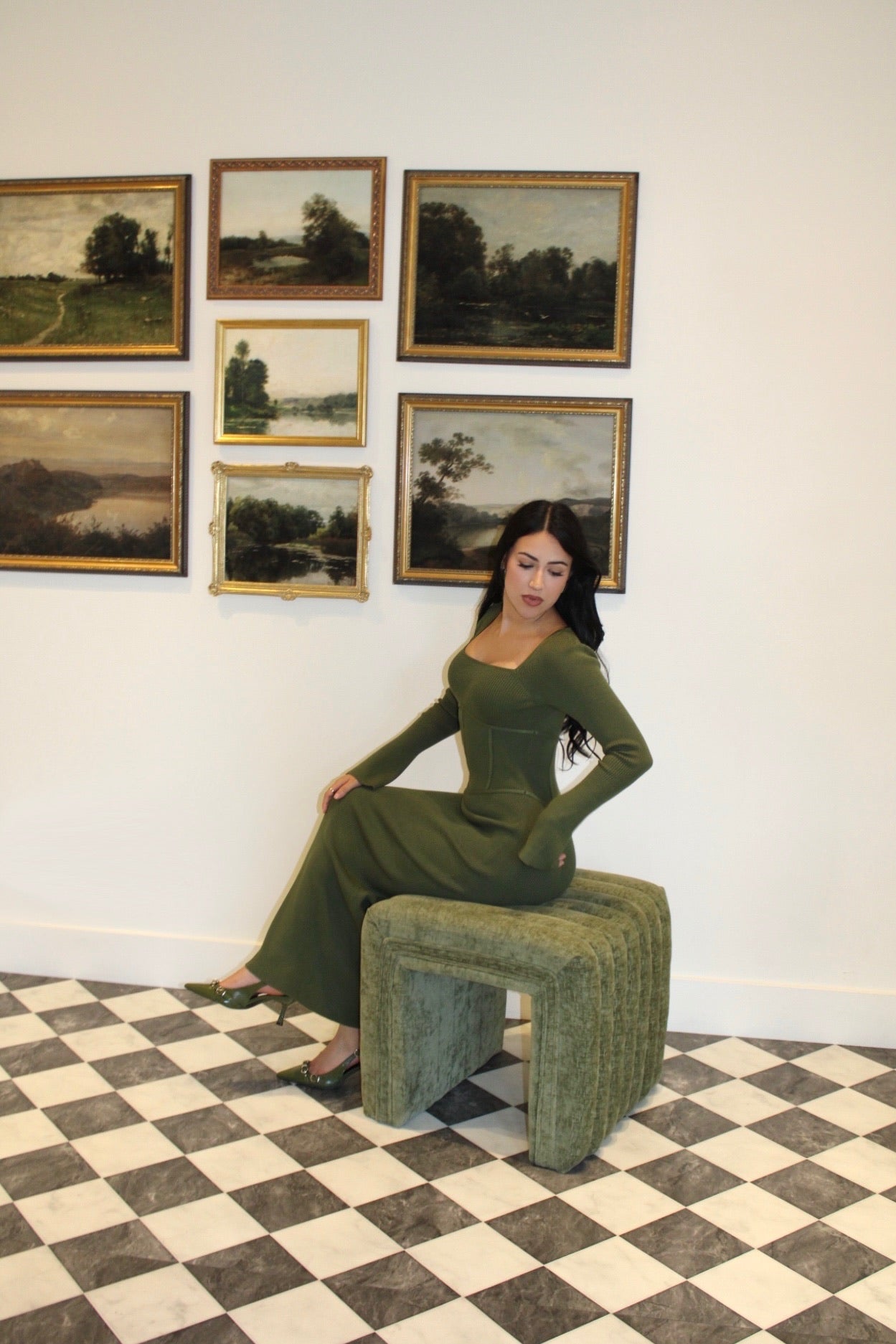 Woman in a green loungewear dress sitting on a green stool in an art gallery with framed paintings on the wall.