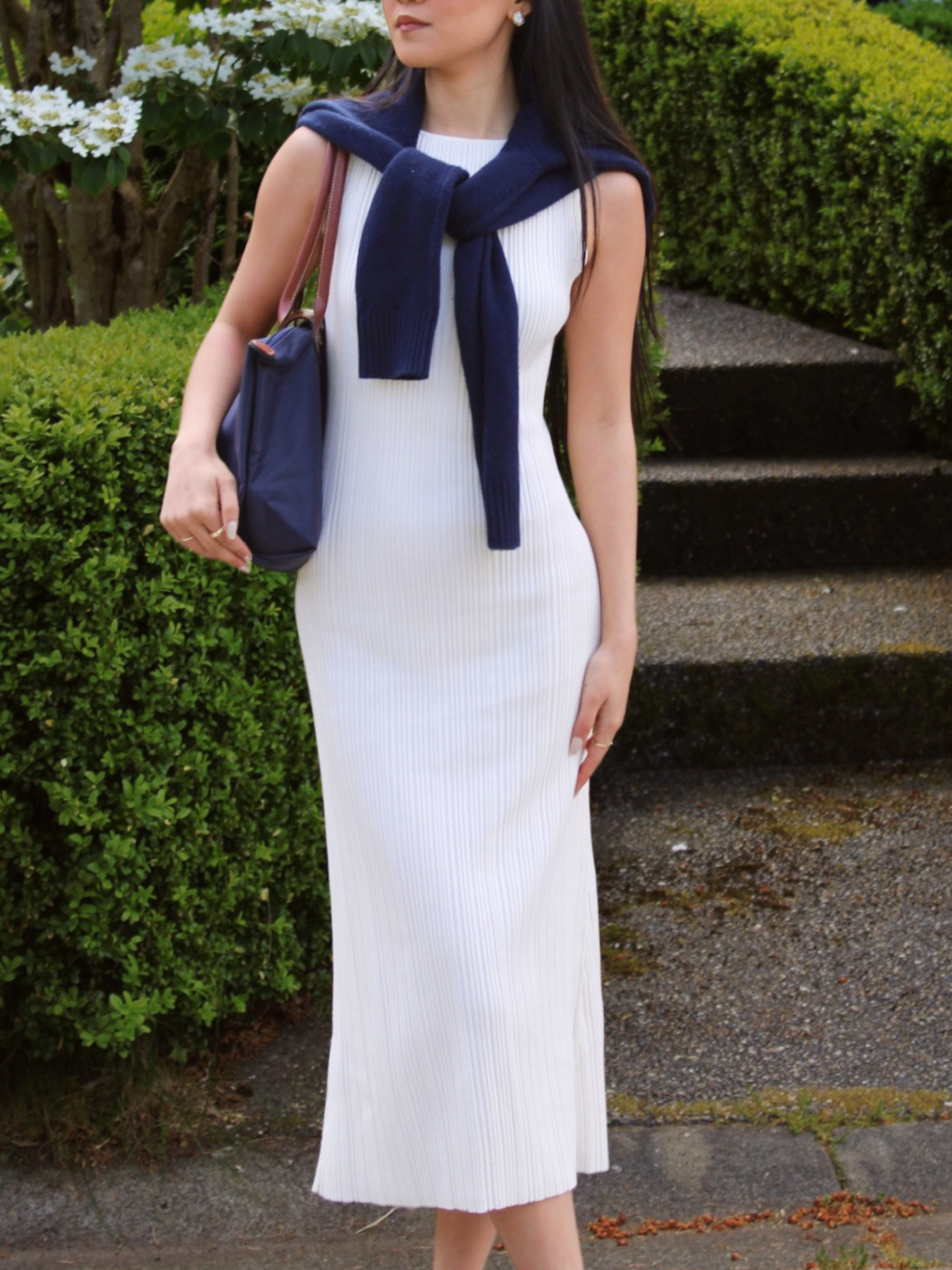 Woman in a white lounge dress with a navy blue cardigan and bag standing outdoors.