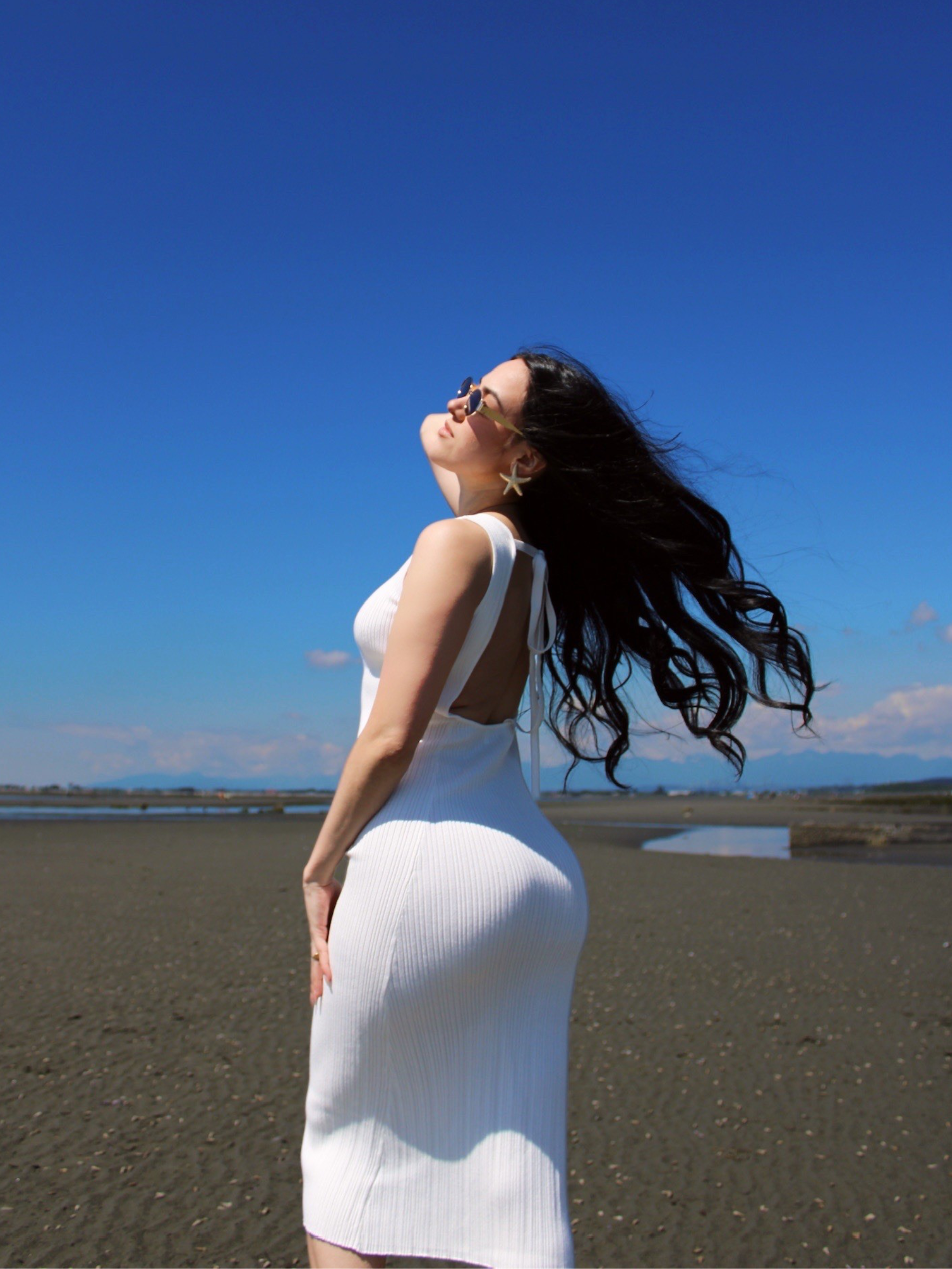 Woman in a white lounge dress standing on a beach with a clear blue sky.